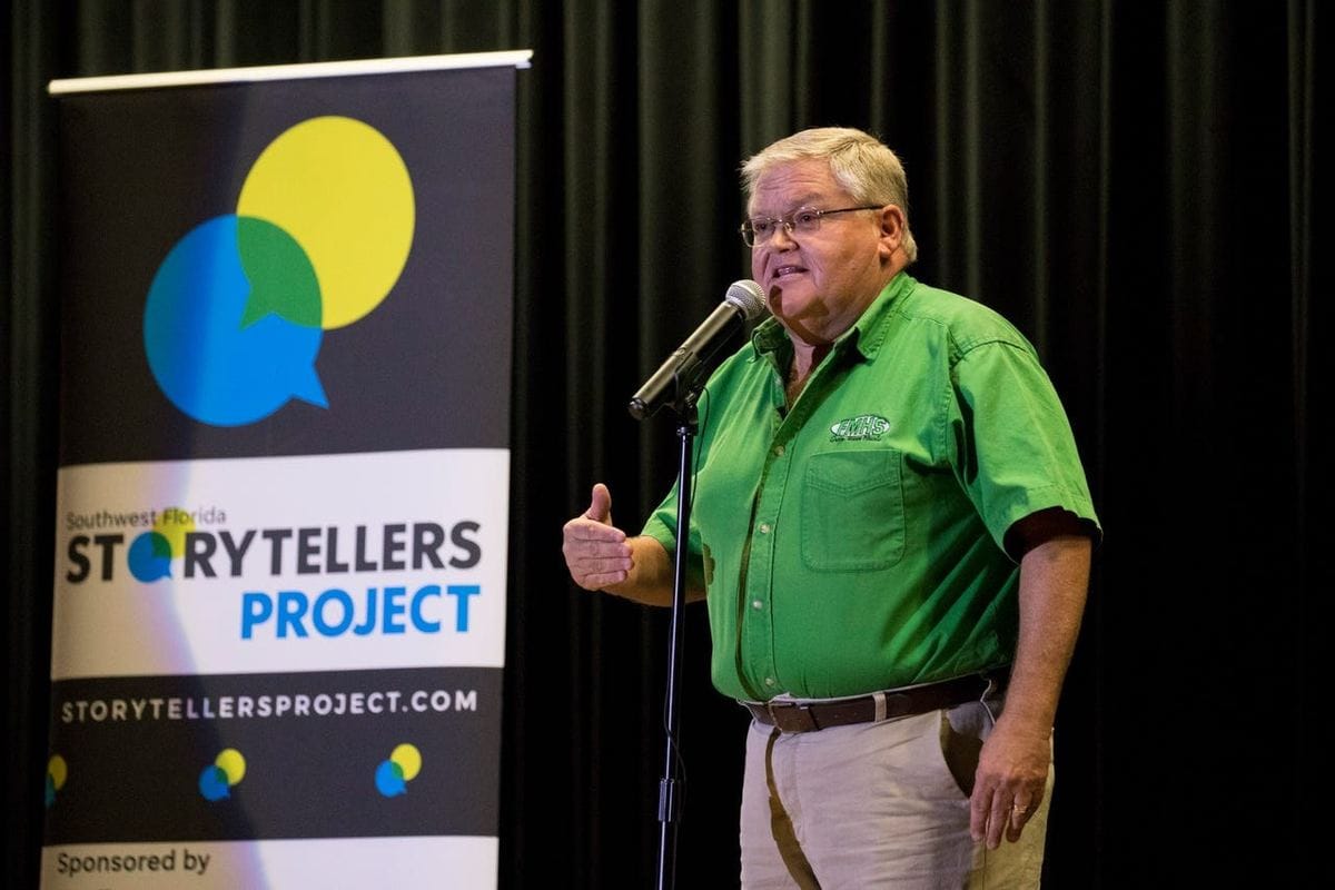 Mark Dahlberg speaking at a microphone at the Southwest Florida Storytellers Project, wearing a Fort Myers High School polo shirt